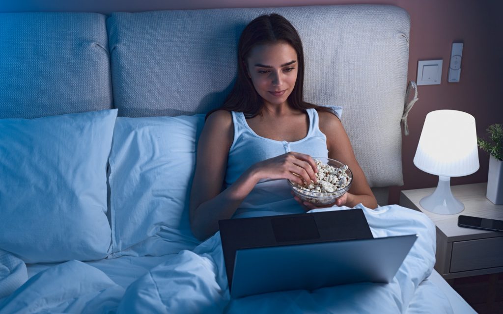guilty pleasures Frau im Bett am Laptop und schaut eine Serie und ist dabei Popkorn